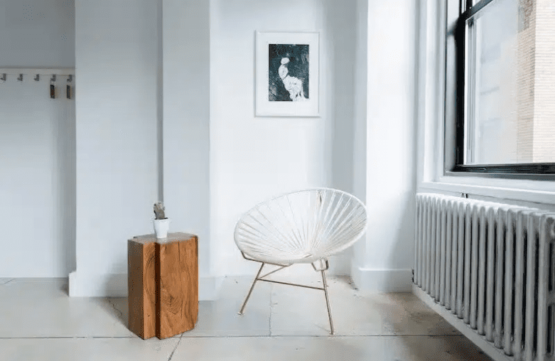 Minimalist white dish chair next to a wooden pedastal with a single plant in a sparse white room.