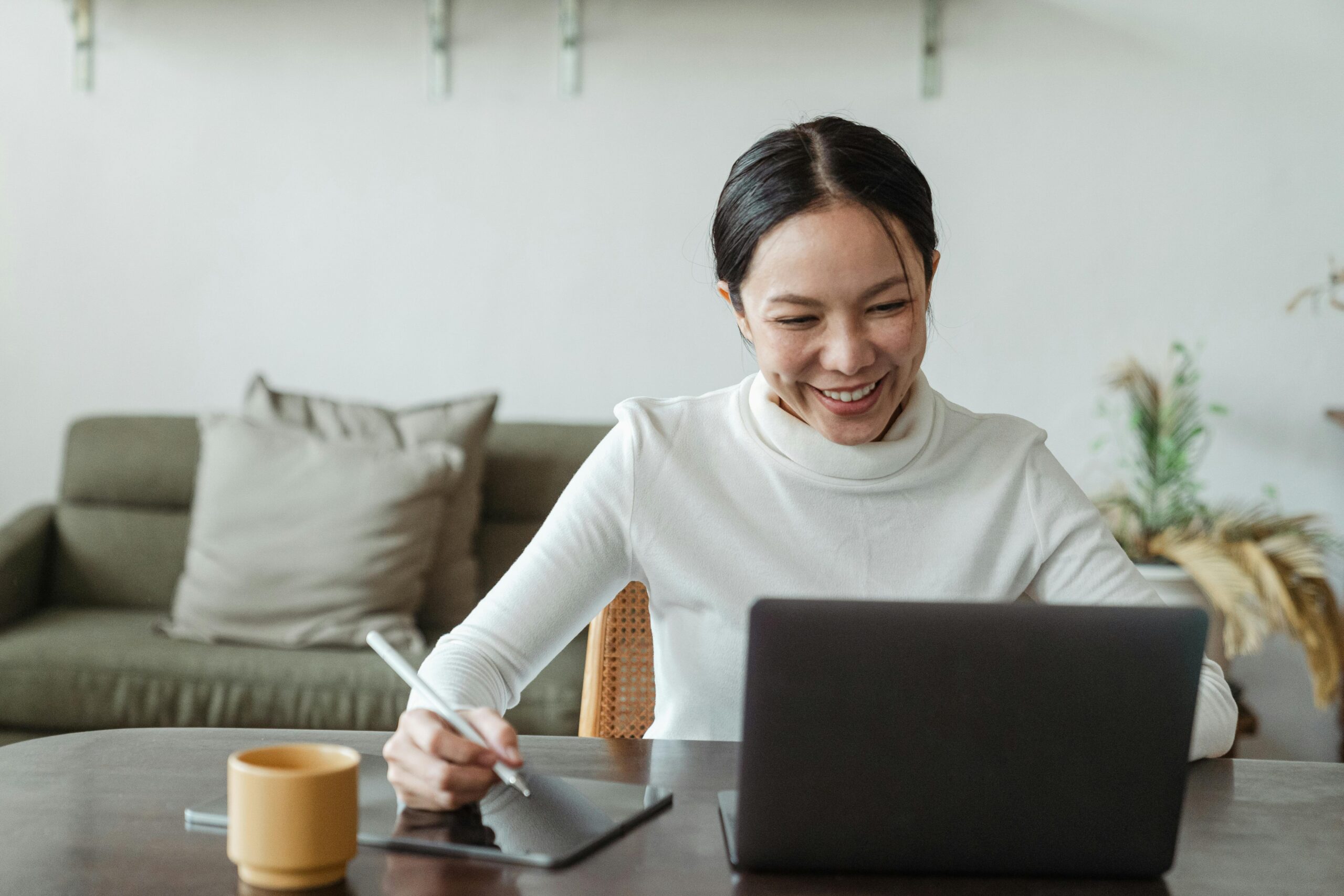 A woman is working remotely. She is smiling and focused, using a stylus on a digital tablet while engaging with her laptop.